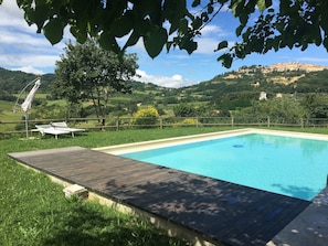 Una piscina rectangular con plataforma de madera, rodeada de abundante vegetación y vistas a un pueblo en la colina.