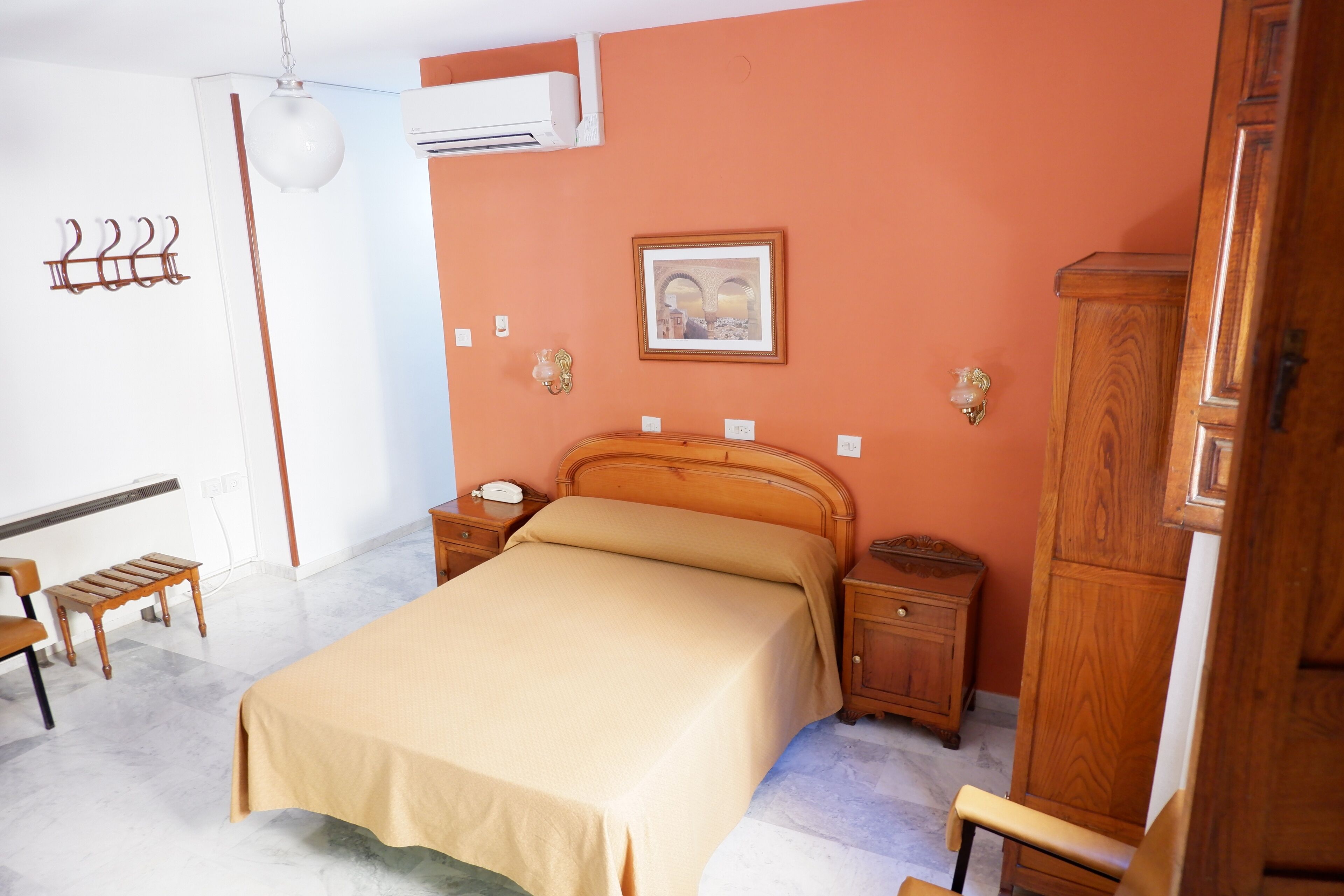 Un dormitorio con cama, mesitas de noche de madera, un armario, un banco, perchero y aire acondicionado instalado en la pared.