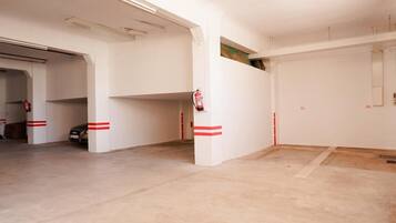 Amplio estacionamiento interior con piso de concreto y paredes blancas, que presenta varios pilares con franjas rojas y un extintor de incendios montado en uno de ellos.