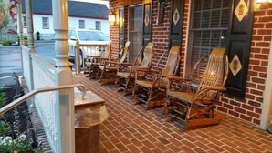 A brick building with a porch featuring wicker rocking chairs and a wooden bench. - Country Living Inn (Lancaster)