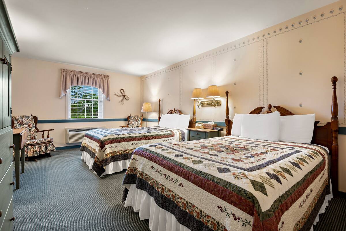 A hotel room with two beds, a desk, a chair, a lamp, and a window with curtains.