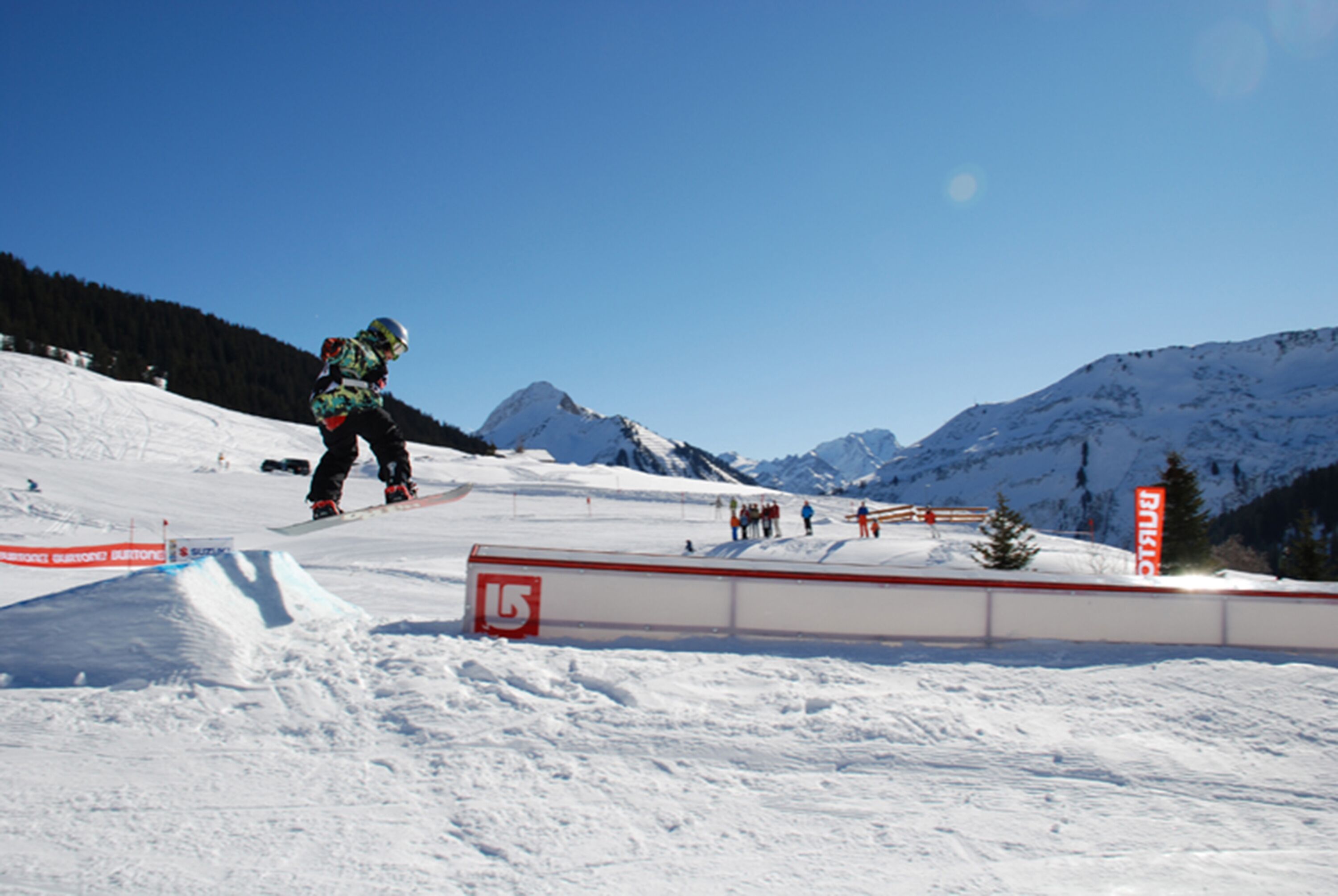 A snowboarder performing a jump on a snowy slope with mountains in the background.