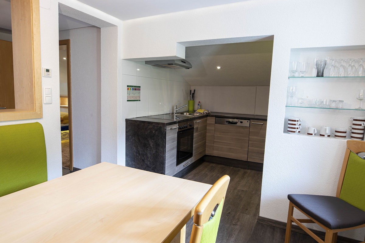 A modern kitchen with a dining area, including a table and chairs, and a glass display shelf with kitchenware.