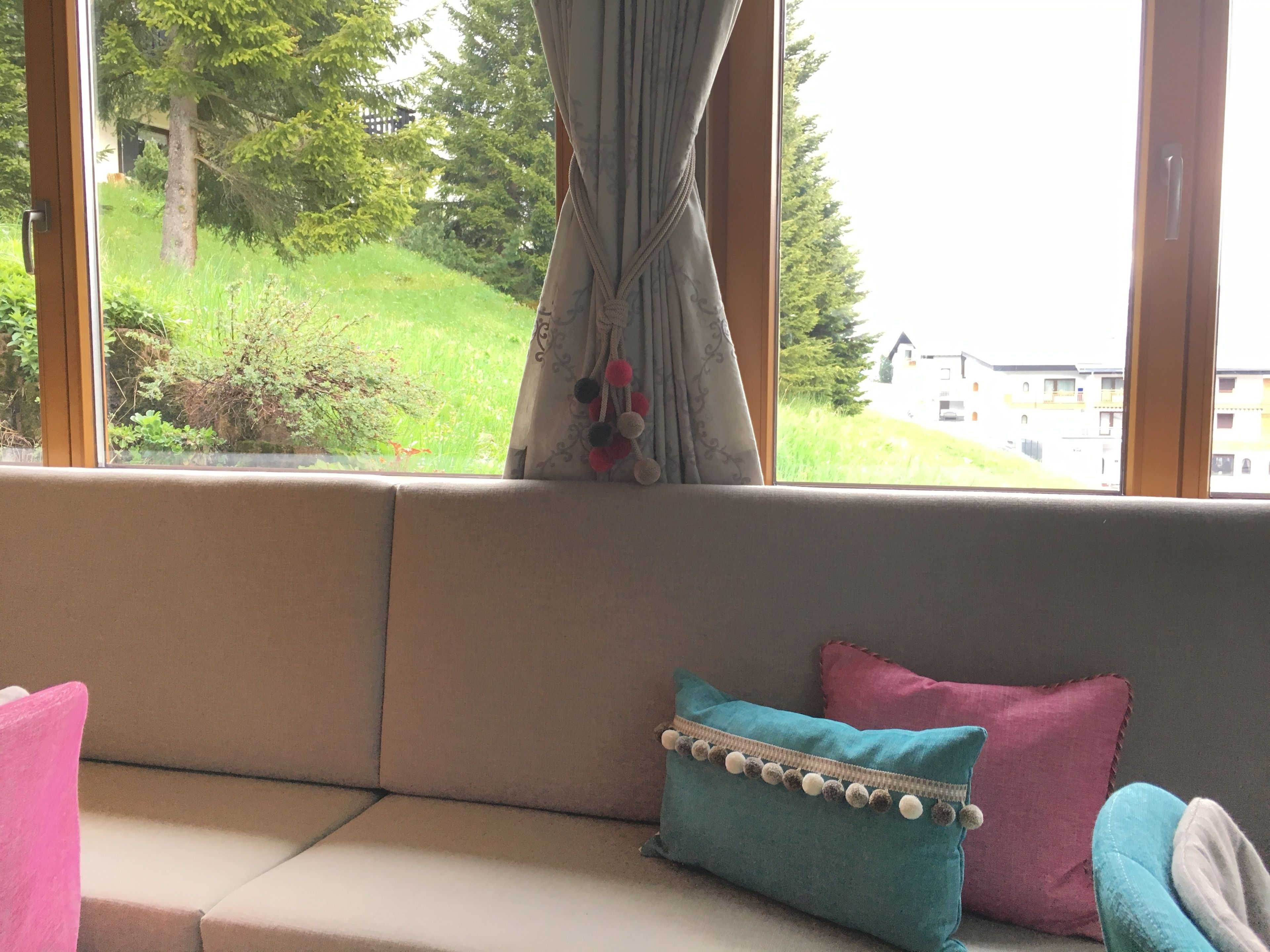 Una ventana con cortina, un sofá con dos almohadones y vistas a zonas verdes y edificios.