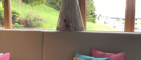 Una ventana con cortina, un sofá con dos almohadones y vistas a zonas verdes y edificios.