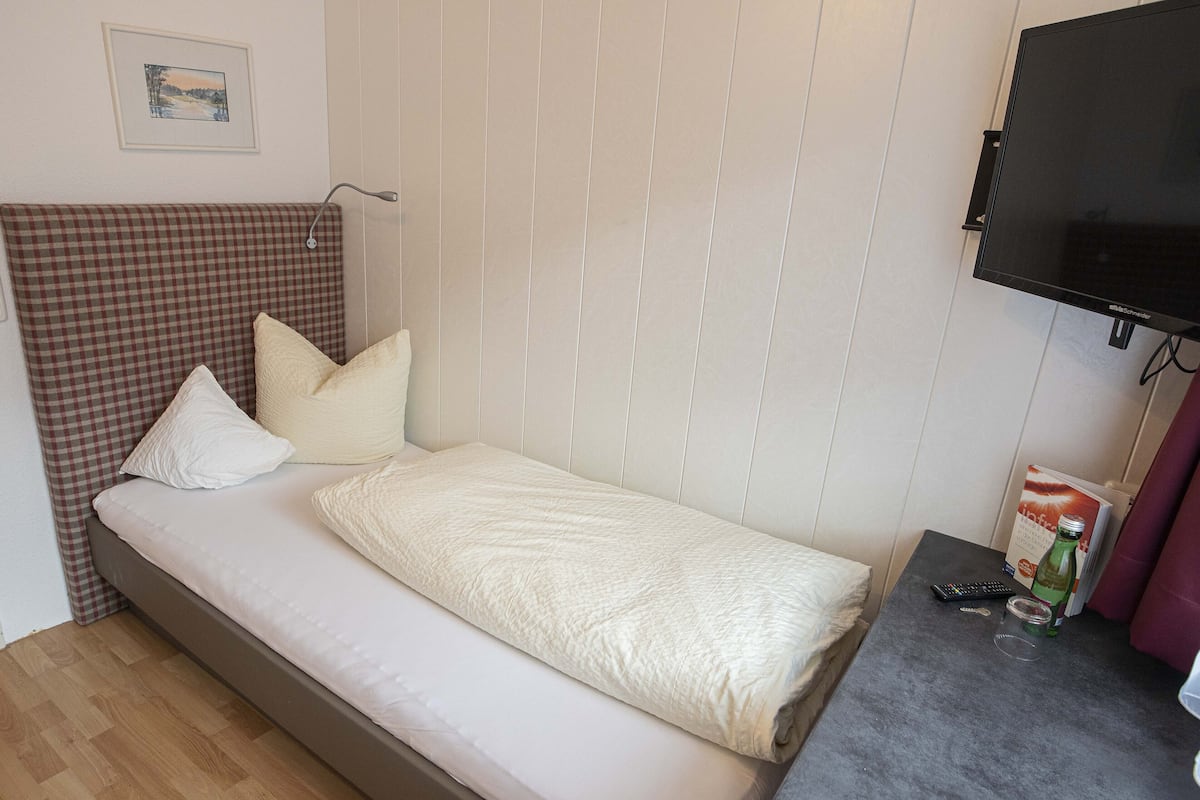 A single bed with a checkered headboard, a flat-screen TV mounted on the wall, and a small table with a remote control and a bottle.