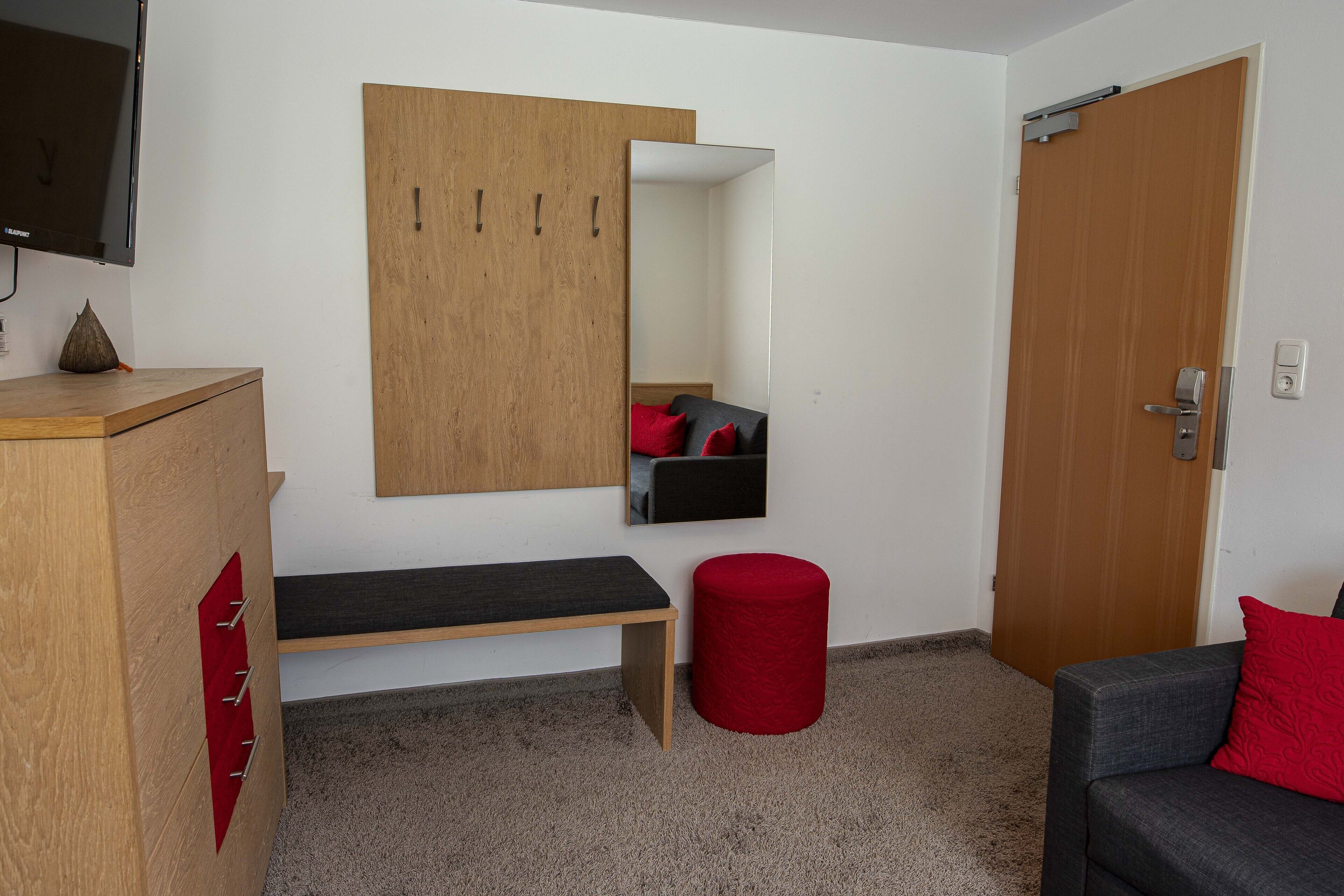 A room with a wooden wardrobe, a bench, a red stool, a grey sofa, a television, and a door.