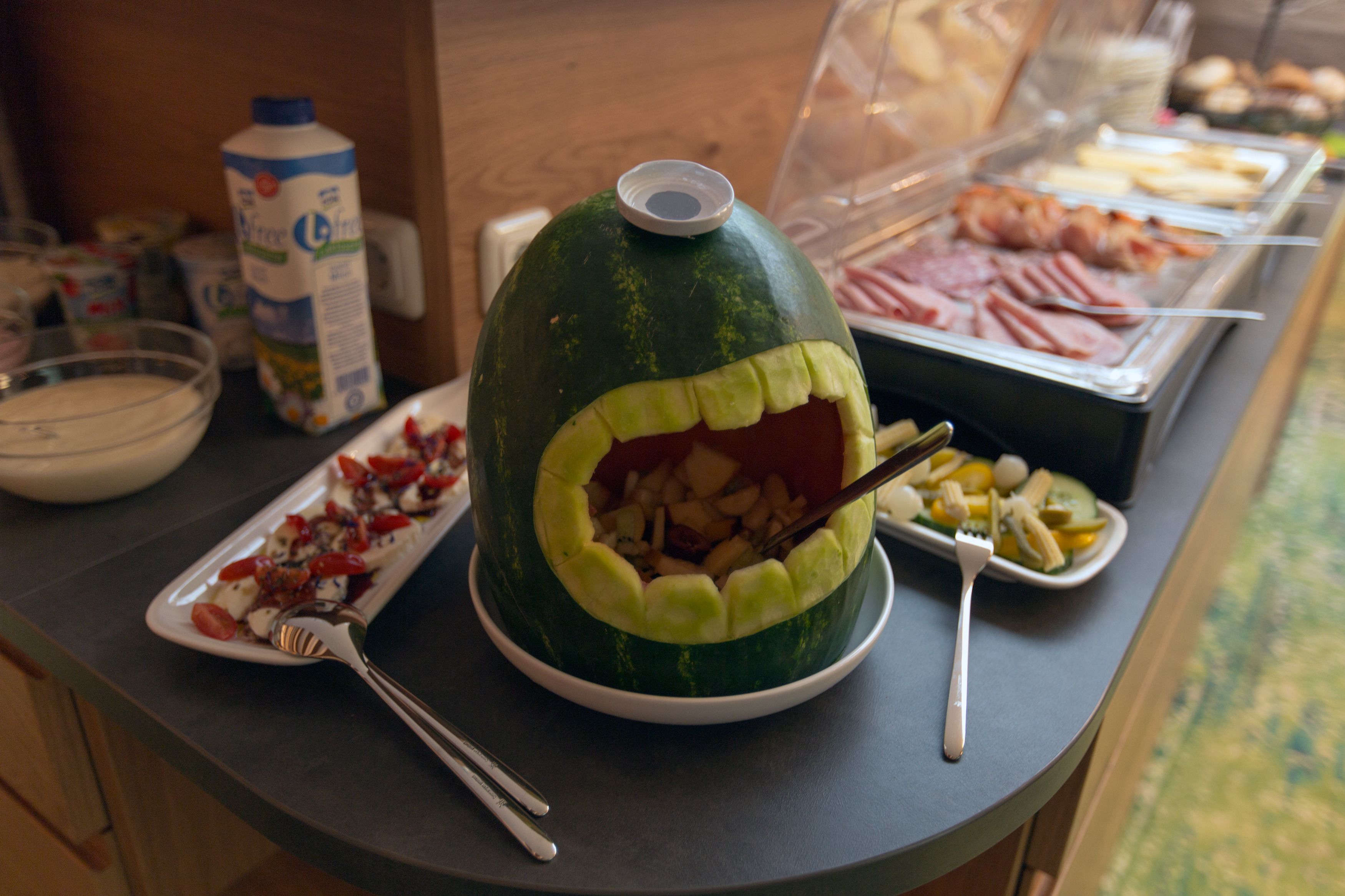 A buffet with a watermelon carved to resemble a face, a milk carton, and various food items.