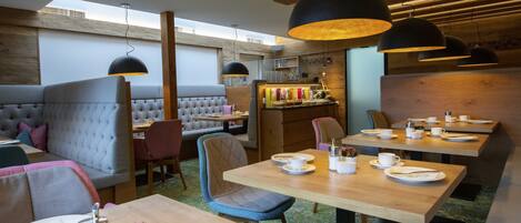 A modern restaurant with wooden tables, cushioned seating, pendant lighting, and a visible bar area.
