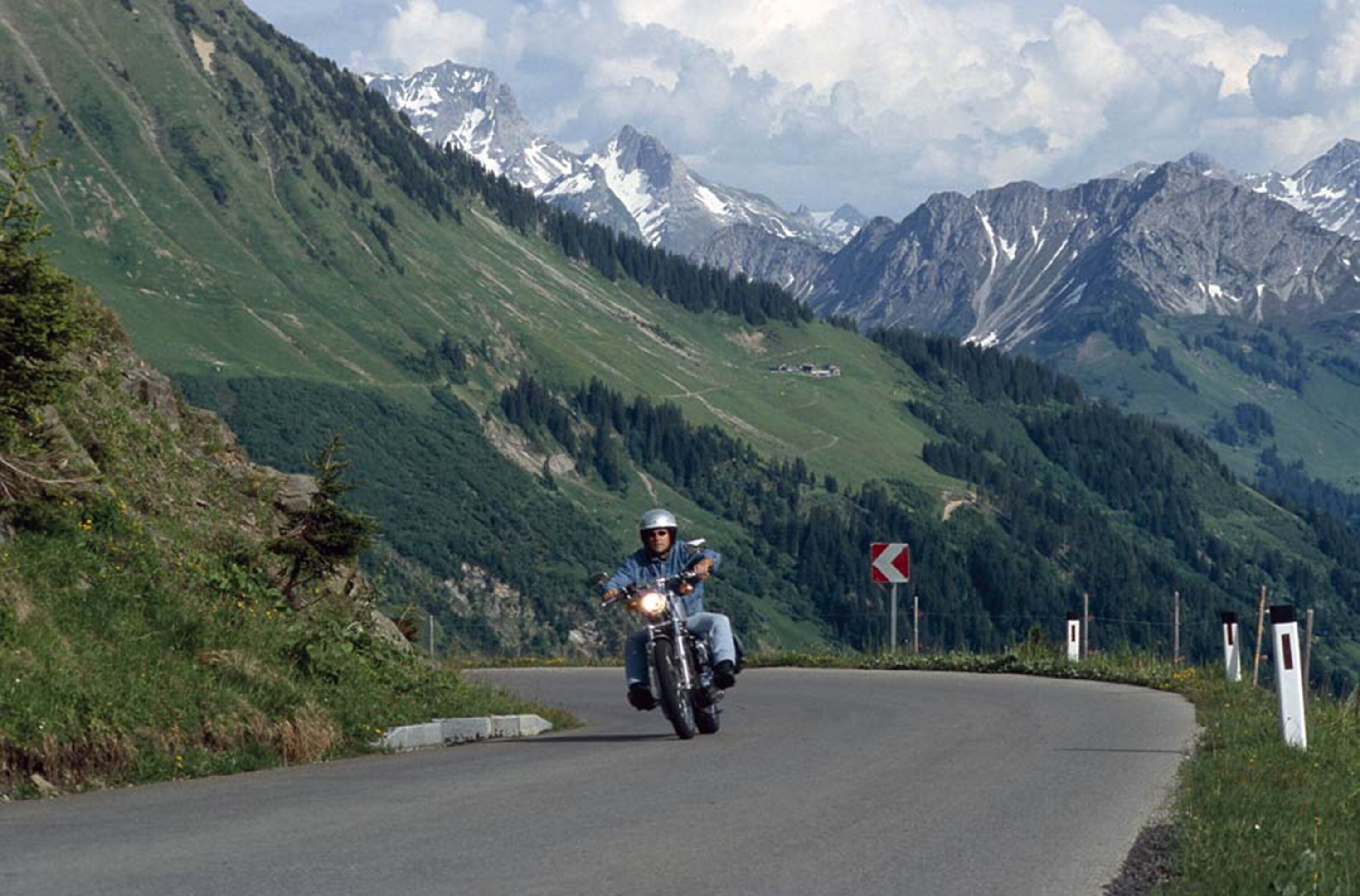 Ein Motorradfahrer auf einer kurvenreichen Bergstraße mit Blick auf schneebedeckte Gipfel und üppige grüne Vegetation.