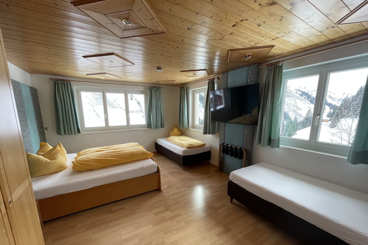 A room with wooden flooring, two beds with yellow bedding, a TV mounted on the wall, and large windows with curtains.