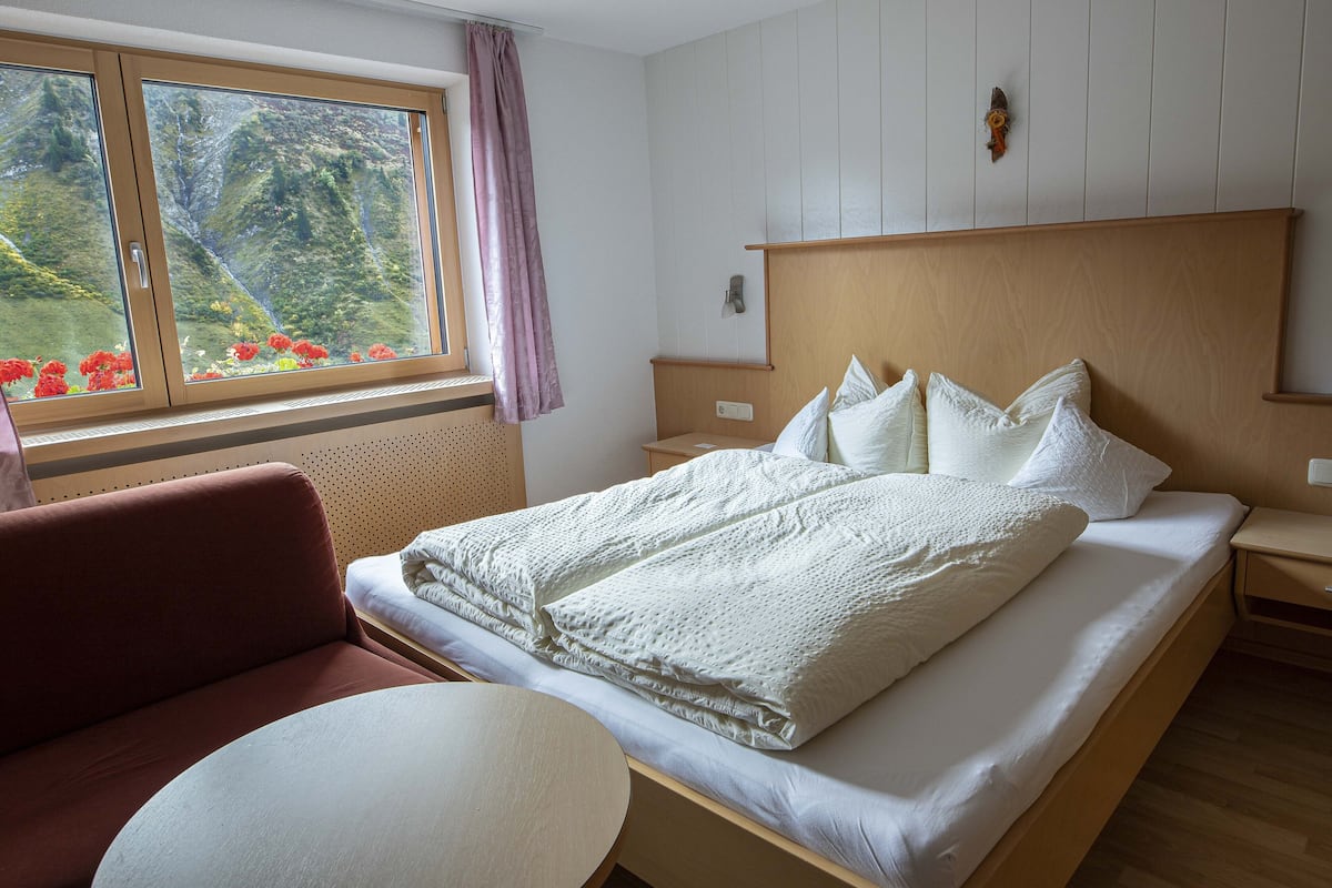 A bedroom with a large bed, a wooden headboard, a bedside table, a round table, and a window with a view of greenery.