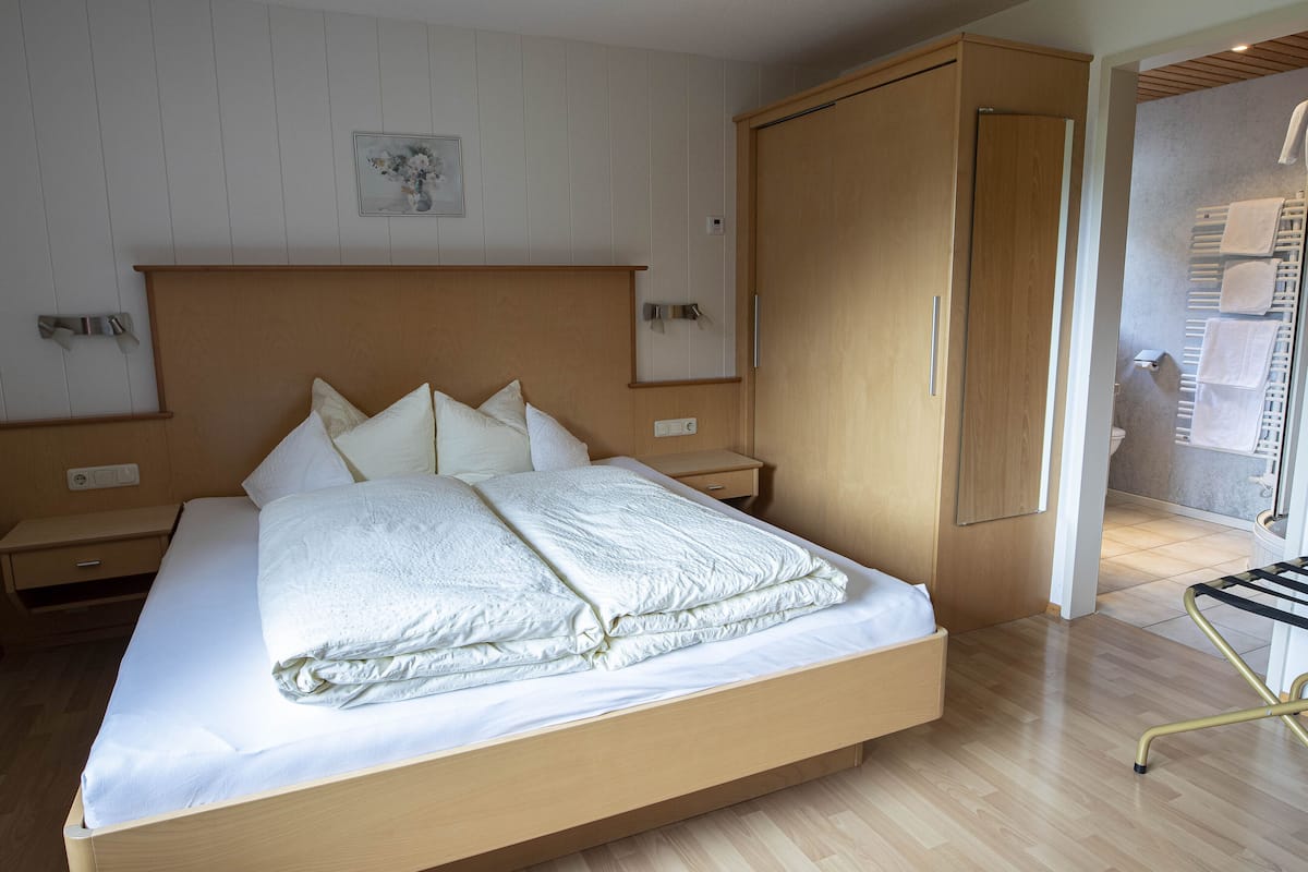 A modern bedroom with a wooden bed, a nightstand, a wardrobe, and a bathroom visible through an open door.