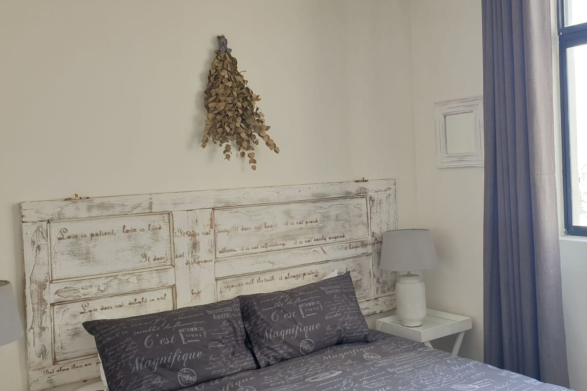 A bedroom with a white wooden headboard, two bedside lamps, a bed with dark patterned bedding, a wall-mounted plant, and a window with a view of buildings.