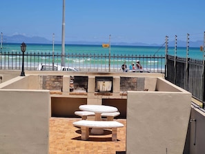 A beachside patio with a stone table and benches, overlooking the ocean and a fenced area with people and vehicles.