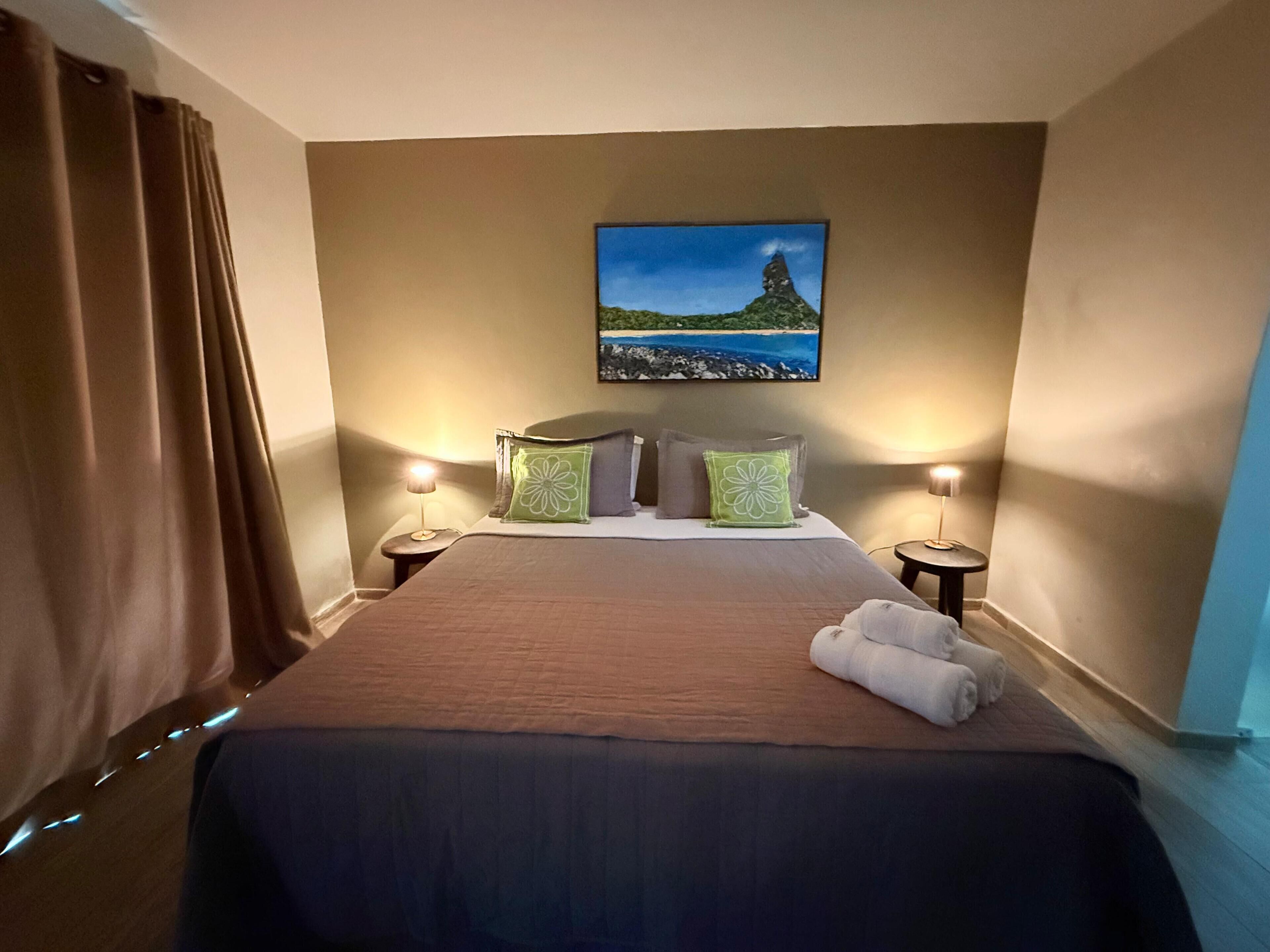 A hotel room with a bed, two bedside lamps, a painting of a landscape, and a window with curtains.