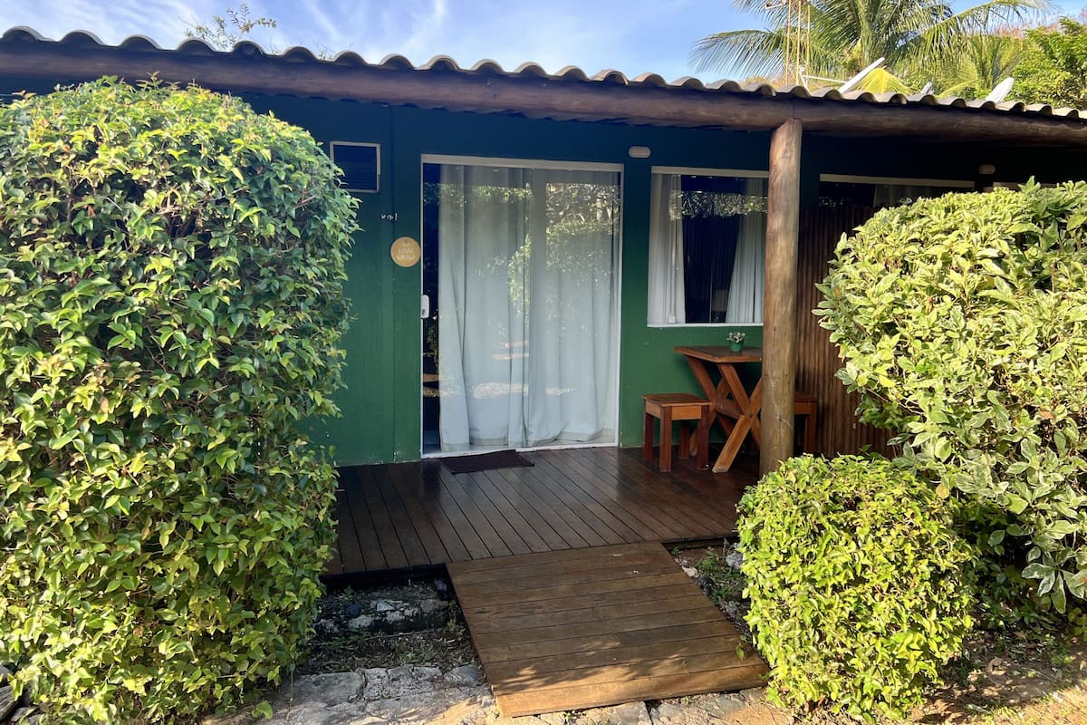 A small, green building with a wooden deck, a window with white curtains, and a small table with a chair.