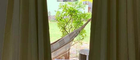 A window with beige curtains, a hammock, and a view of a green lawn and a building.