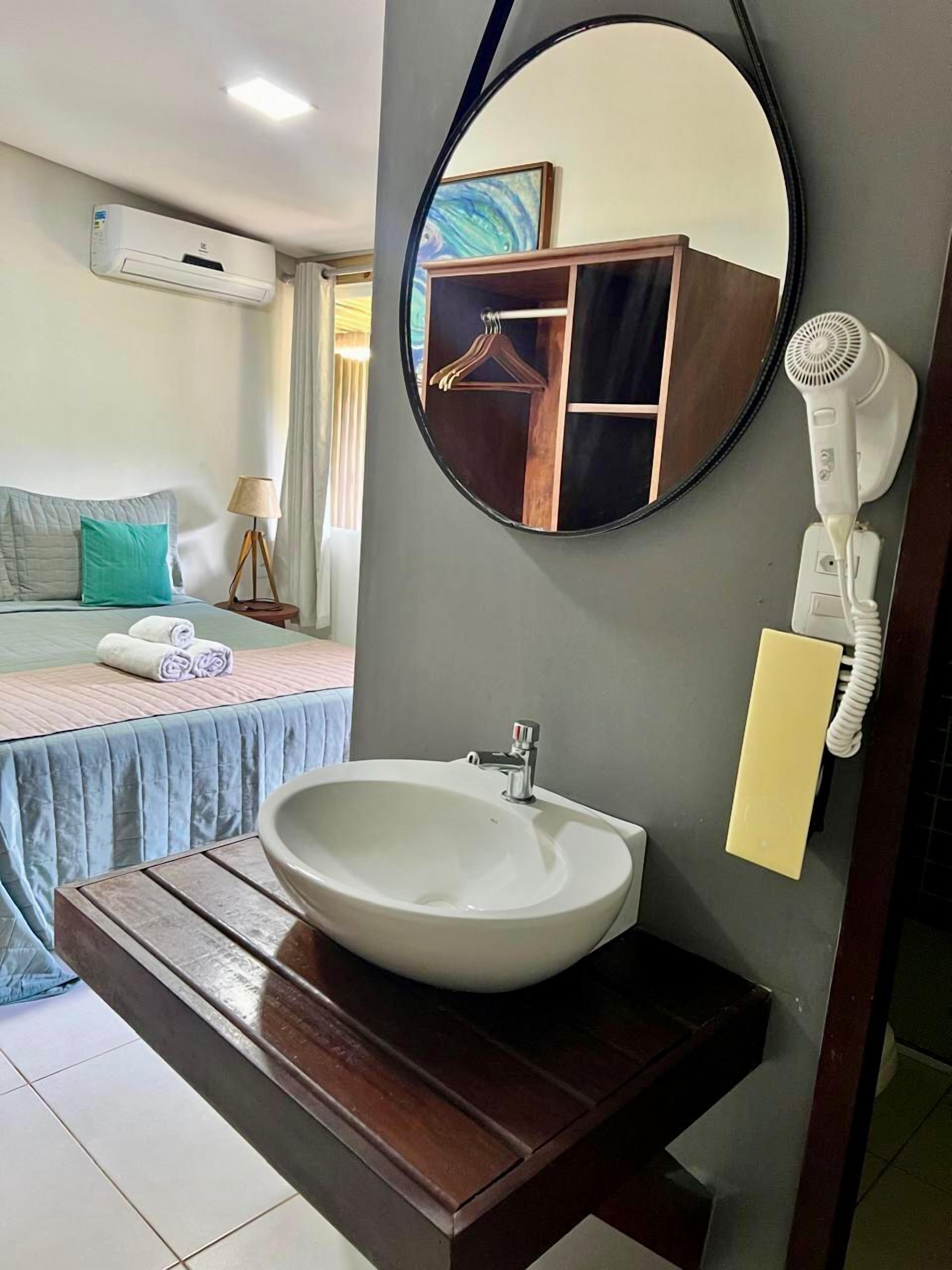 A hotel room with a bed, a bedside table, a round mirror, a white sink, and a hairdryer.