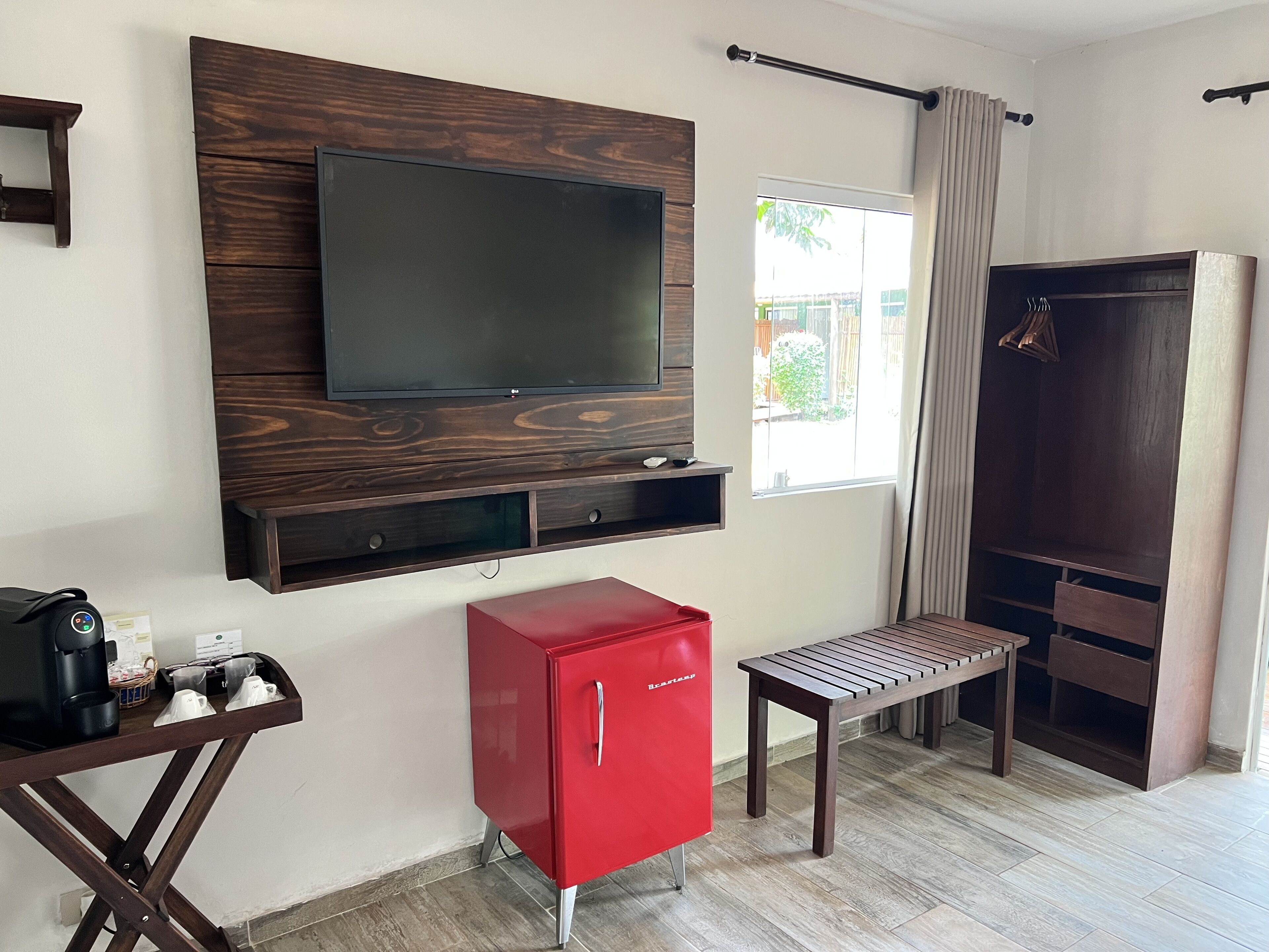 A room with a mounted TV, a red minibar, a wooden bench, and a wardrobe.