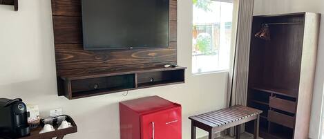 A room with a mounted TV, a red minibar, a wooden bench, and a wardrobe.