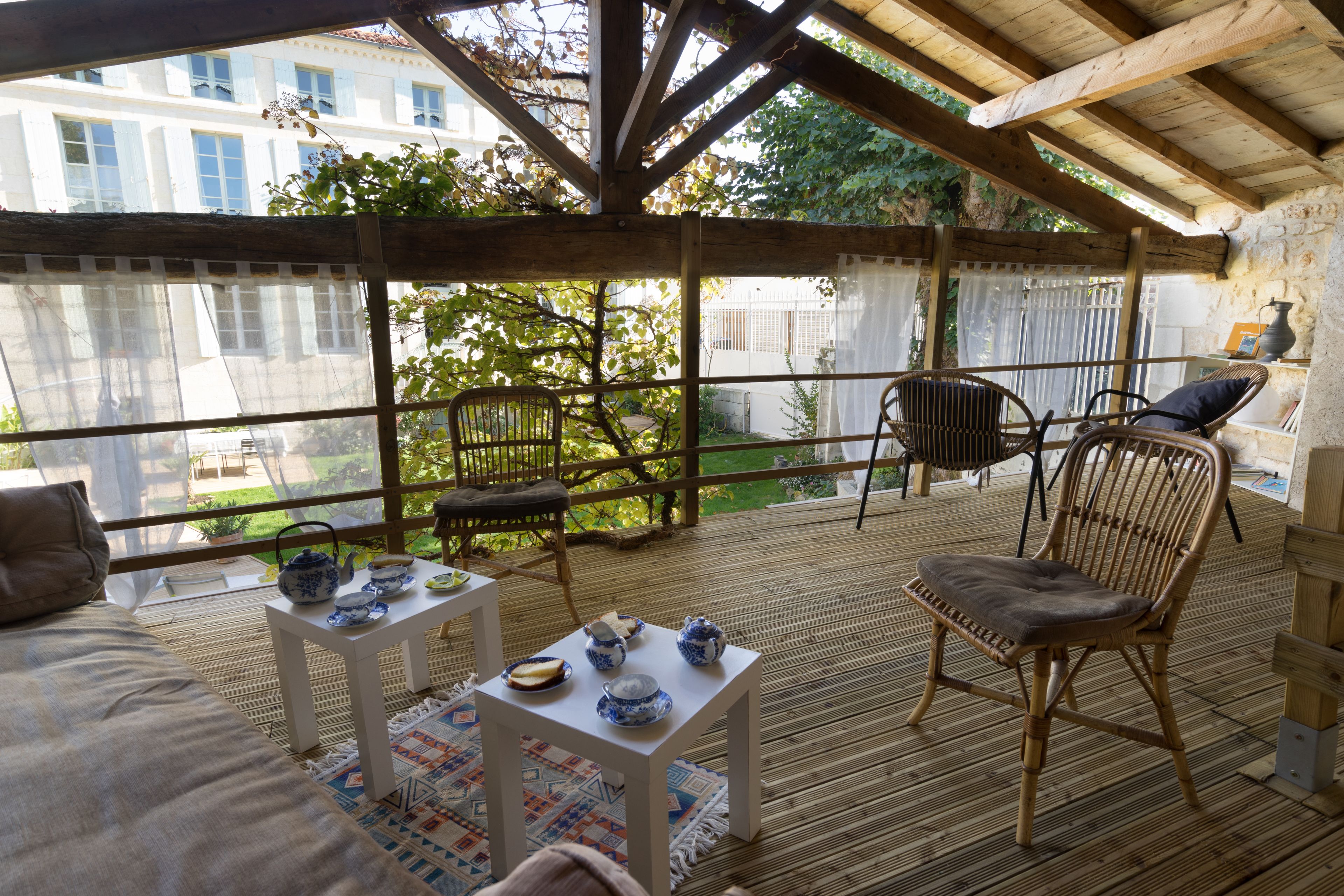 Un espacio al aire libre techado con piso de madera, sillas de mimbre, una pequeña mesa con objetos de cerámica azul y blanca, y vistas a un jardín y edificios al fondo.