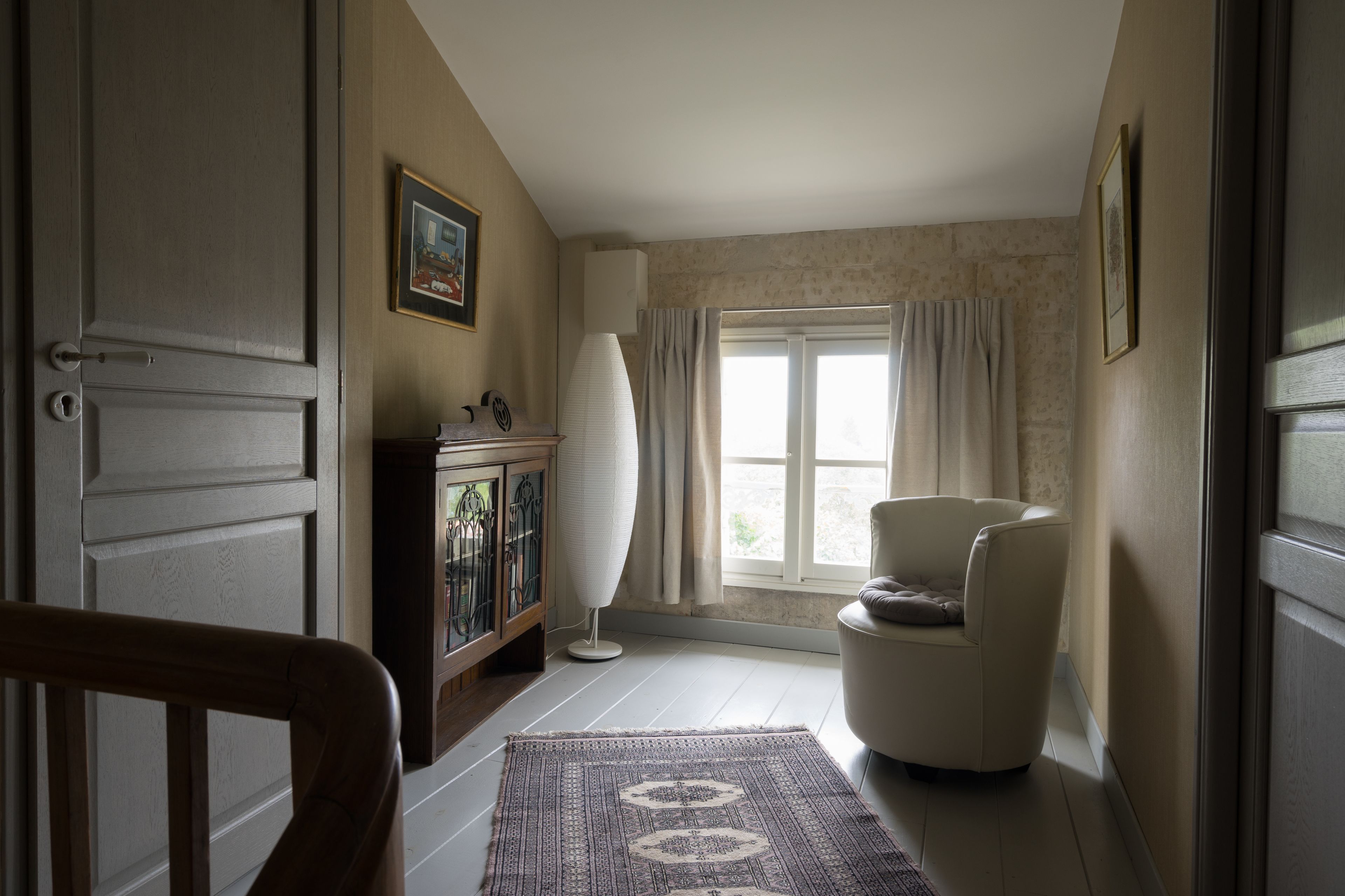 A hallway with a patterned rug, a white chair, a cabinet with glass doors, and a window with curtains.