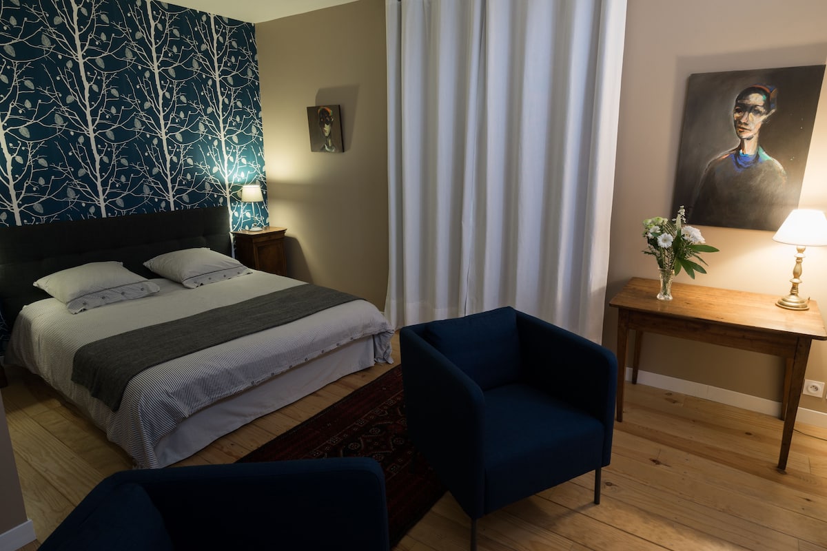 Une chambre d’hôtel bien rangée, avec un lit, deux chaises, une table avec une lampe et un tableau accroché au mur.