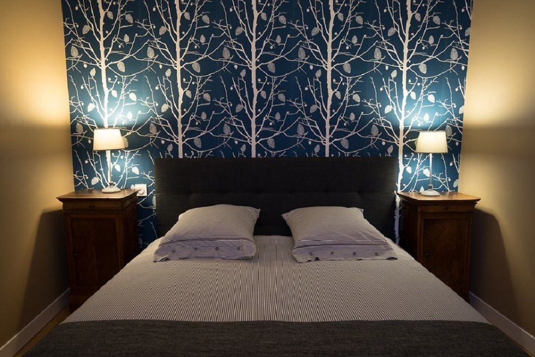 A neatly made bed with a striped bedspread and two pillows, flanked by two bedside tables with lamps, against a wall with a tree pattern wallpaper.