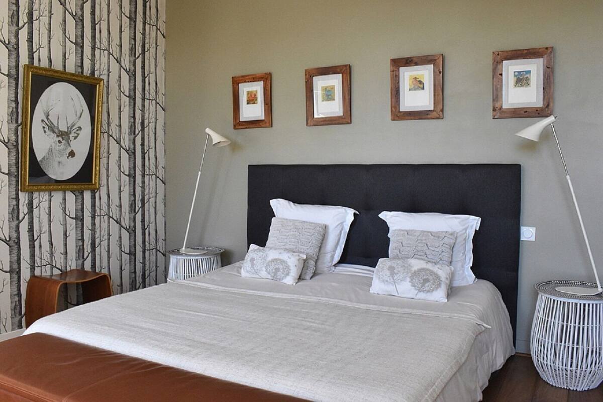 Un lit bien fait, avec une tête de lit sombre, une literie blanche et plusieurs oreillers. Des tableaux encadrés sont accrochés au mur au-dessus du lit.