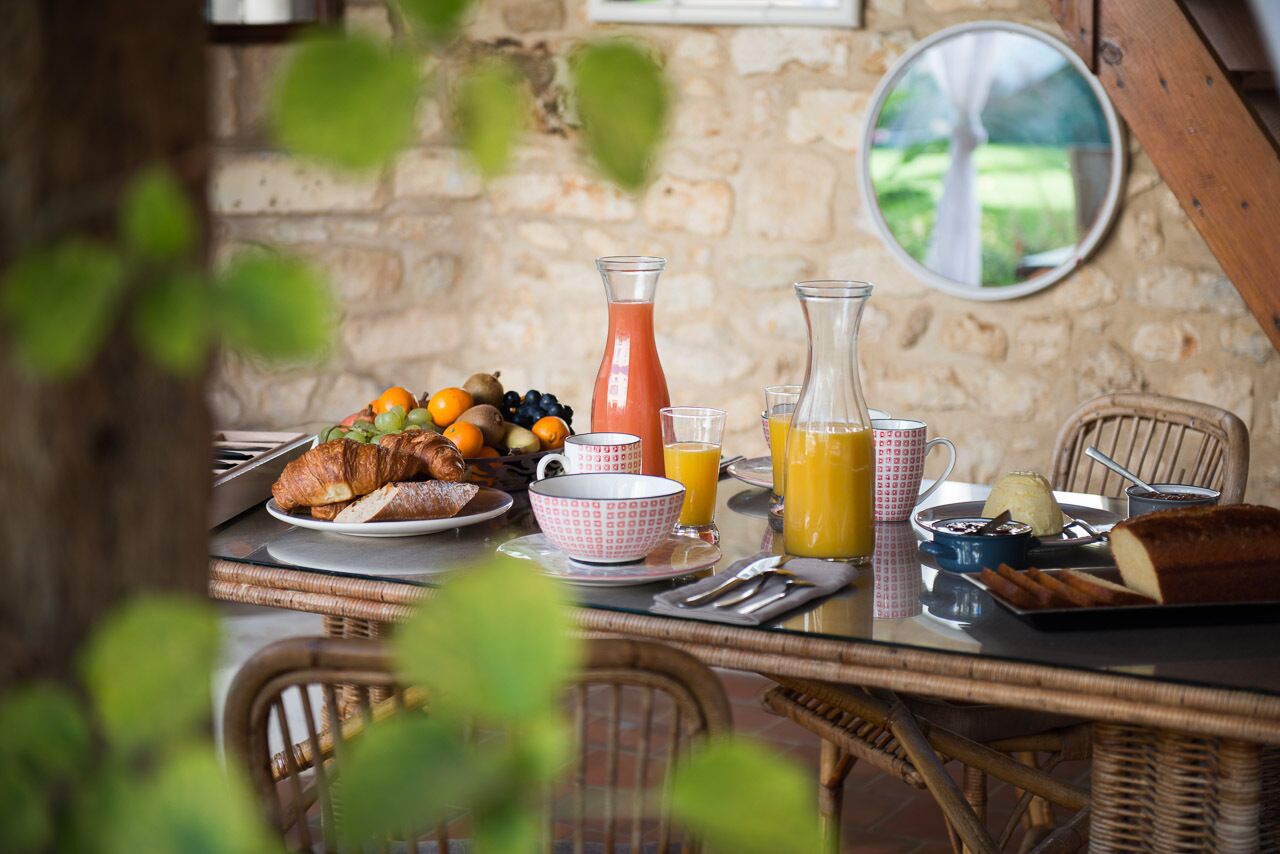 A breakfast setting with croissants, fruits, juice, and a bottle of juice on a table with wicker chairs.