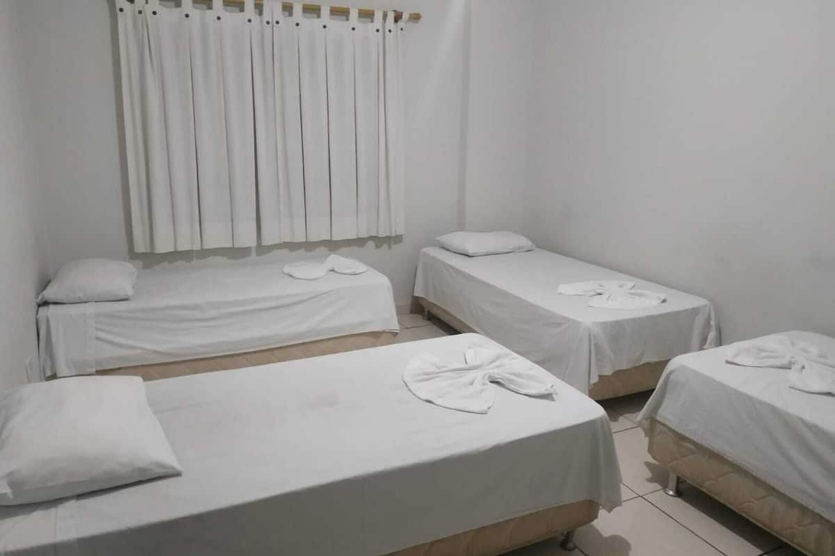 A room with three single beds, each with a white bedspread and a towel on the bed. There is a curtain on the window, and the walls are painted white.