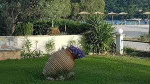 A garden area with a stone wall, a large decorative planter, and a view of a beach with umbrellas. - Marathon Studios &Apartments (Zakynthos)