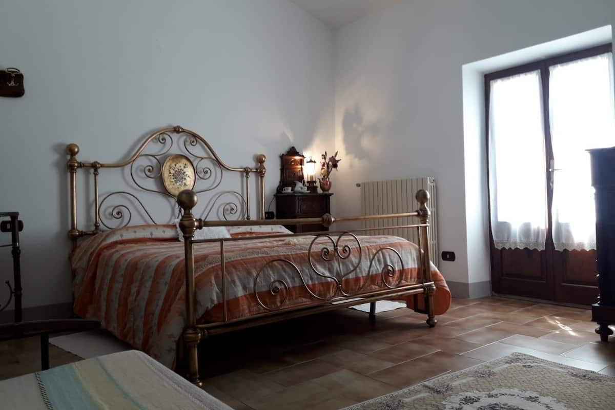Une chambre à coucher avec un lit à ossature métallique, une table de chevet avec une lampe, un radiateur et une fenêtre avec des rideaux.