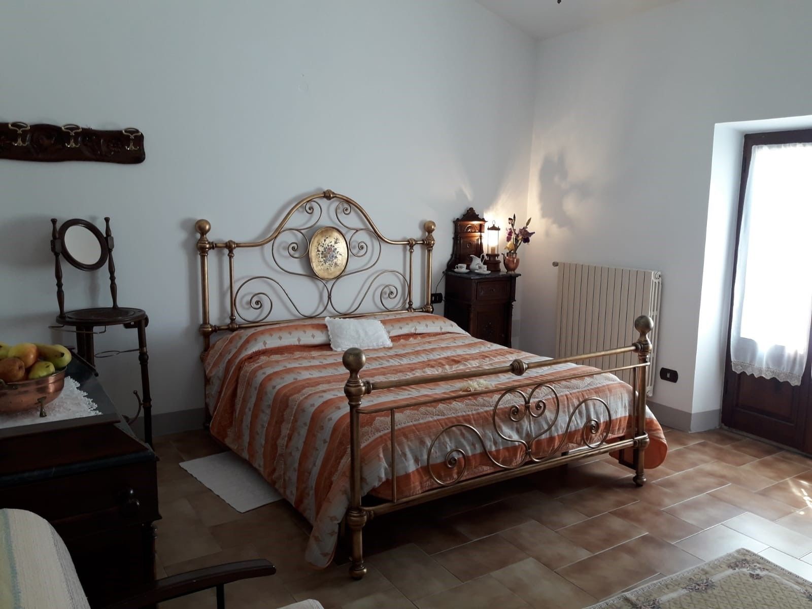 Un dormitorio con una cama de estructura metálica, un aparador de madera, una silla y una mesita con un cuenco de fruta.