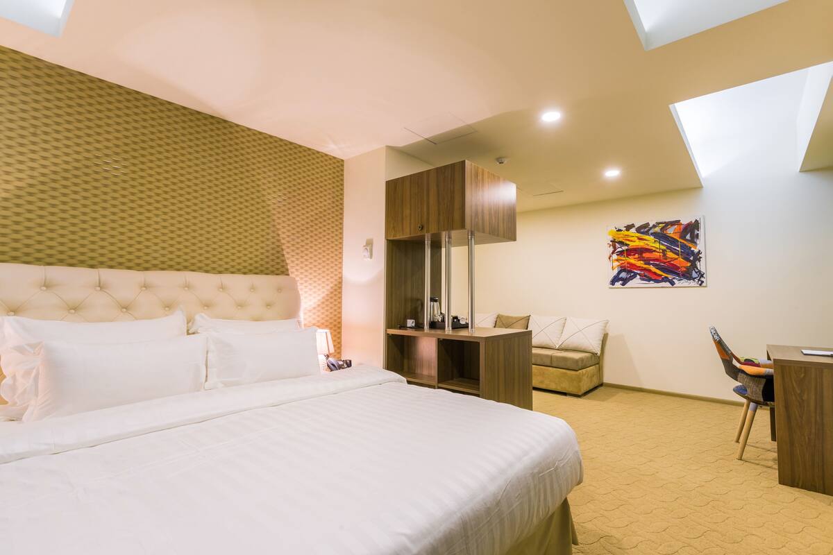 Une chambre d’hôtel avec un grand lit, une armoire en bois, un bureau et une peinture abstraite aux couleurs vives accrochée au mur.