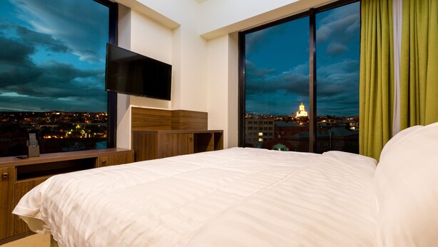 A hotel room with a large bed, a TV mounted on the wall, and a view of a cityscape at dusk. - Pirimze Plaza (Tbilisi)