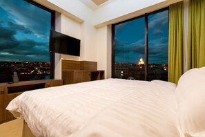A hotel room with a large bed, a TV mounted on the wall, and a view of a cityscape at dusk. - Pirimze Plaza (Tbilisi)
