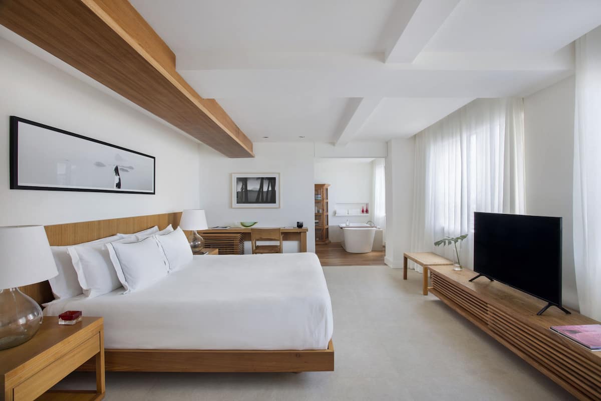 A modern hotel room with a large bed, bedside tables, a TV, and a bathroom visible through an open door.