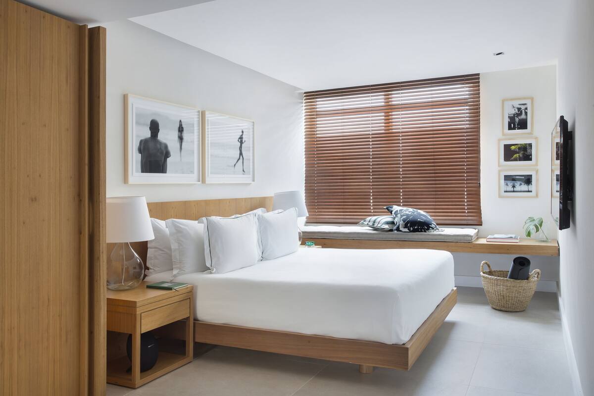 A modern bedroom with a wooden bed, white bedding, a bedside table with a lamp, a window with blinds, and framed pictures on the wall.