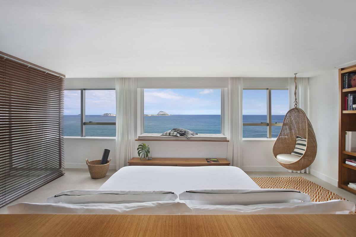 A bedroom with a large bed, a hanging chair, a window seat with a view of the sea, and a bookshelf.