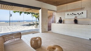 A modern reception area with a wooden ceiling, large windows offering a view of the beach, and a reception desk with a framed artwork of palm trees. - JANEIRO Hotel (Rio de Janeiro)