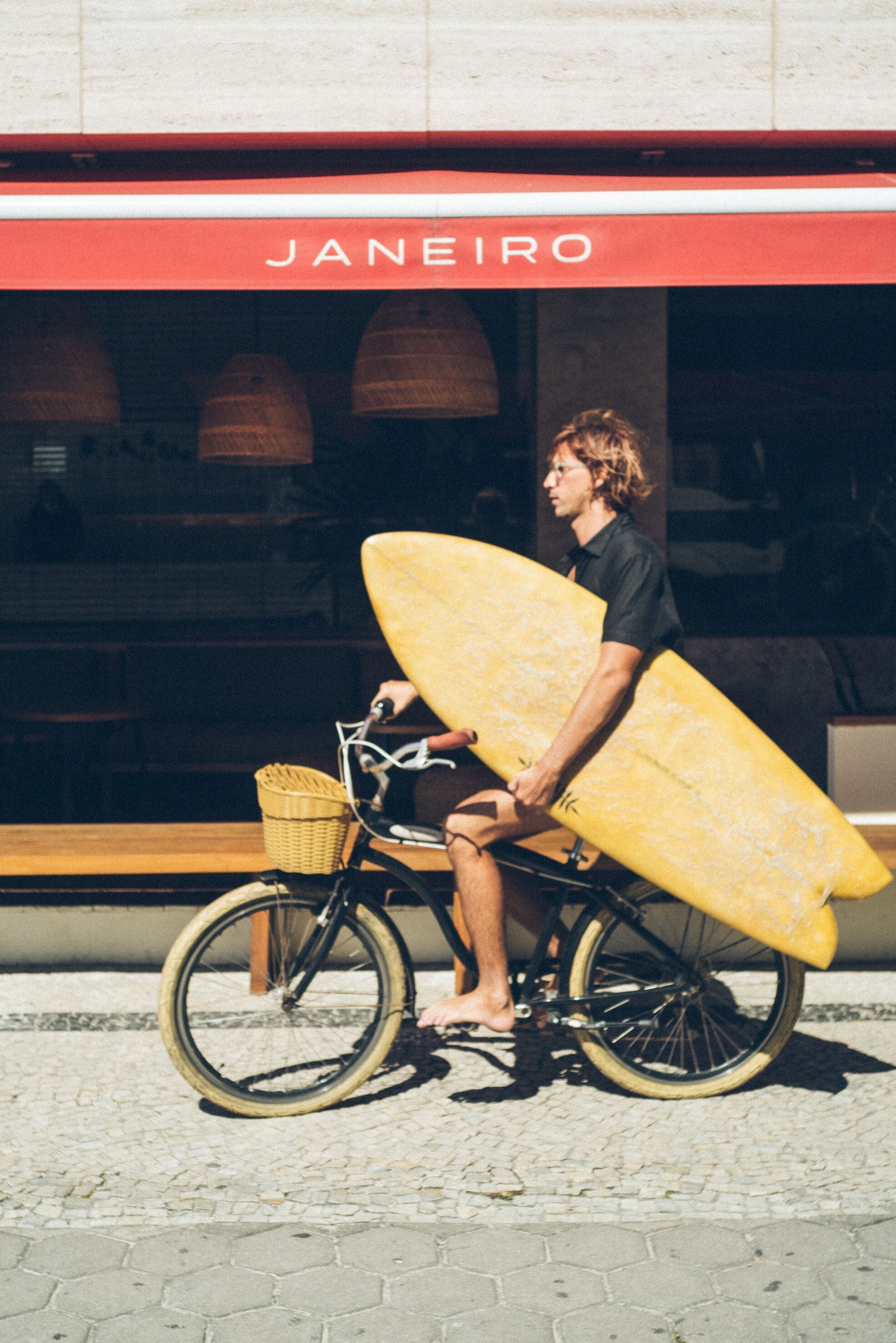 A person on a bicycle with a surfboard, in front of a building with a red awning labeled "JANEIRO".