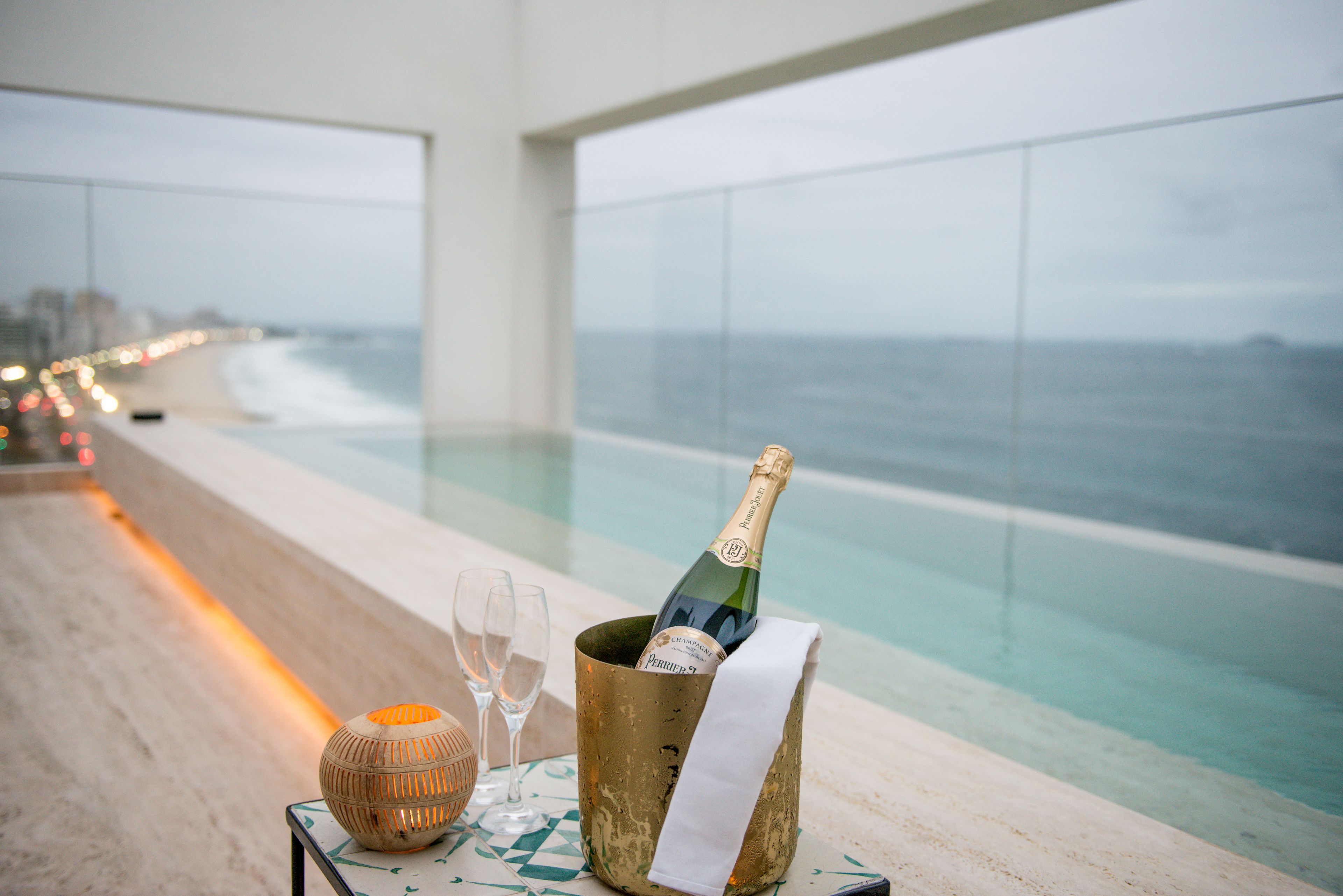 Una bottiglia di champagne in un secchio dorato con un tovagliolo bianco, due bicchieri e una candela su un tavolo vicino a una piscina con vista sulla spiaggia e sull'oceano.