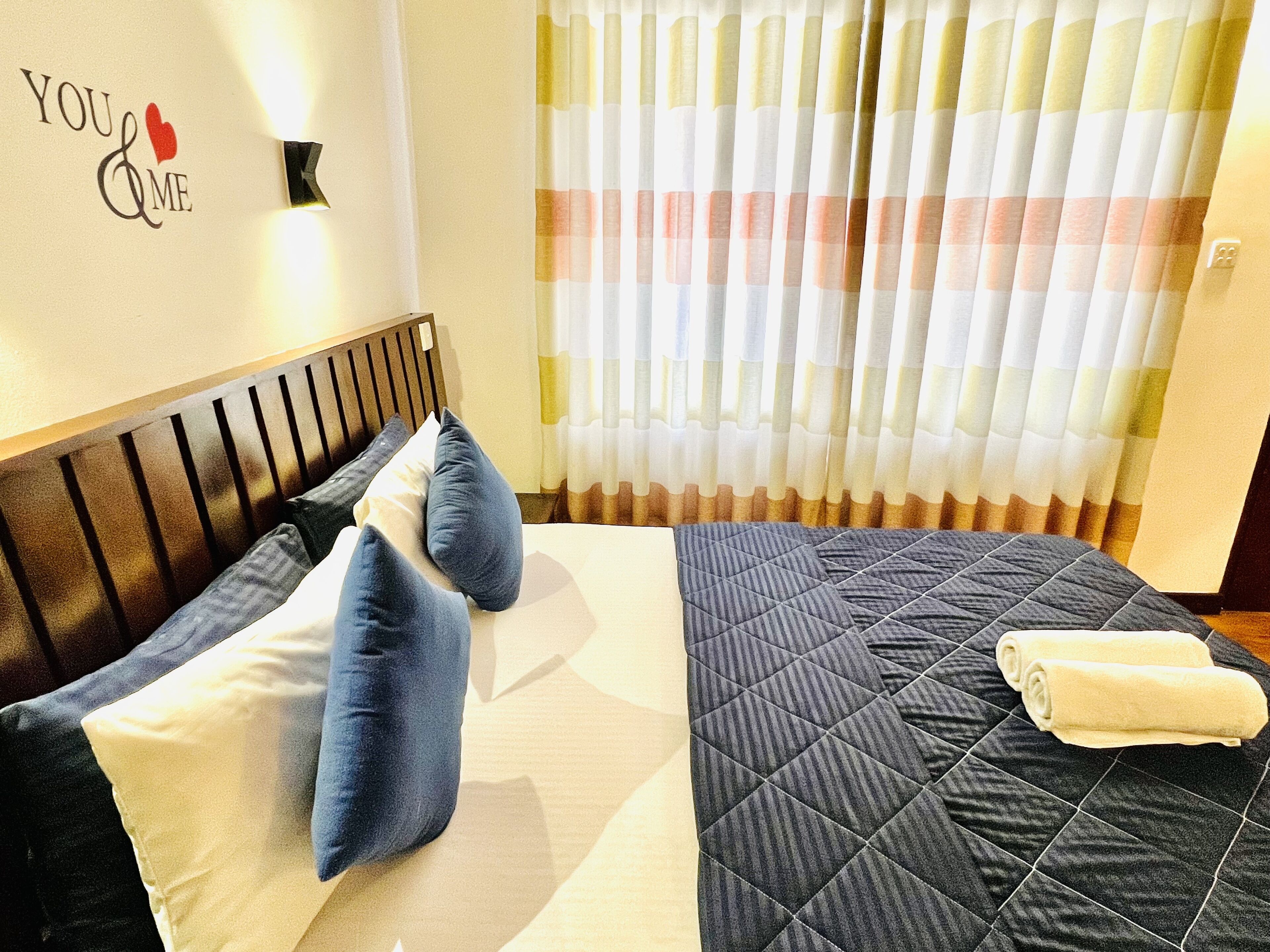 A hotel room with a bed, pillows, a quilted bedspread, a towel, a wall with a 'You & Me' sign, and a window with curtains.