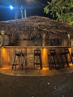 Un área de estar rústica al aire libre con techo de paja, barra de madera y tres taburetes de bar.