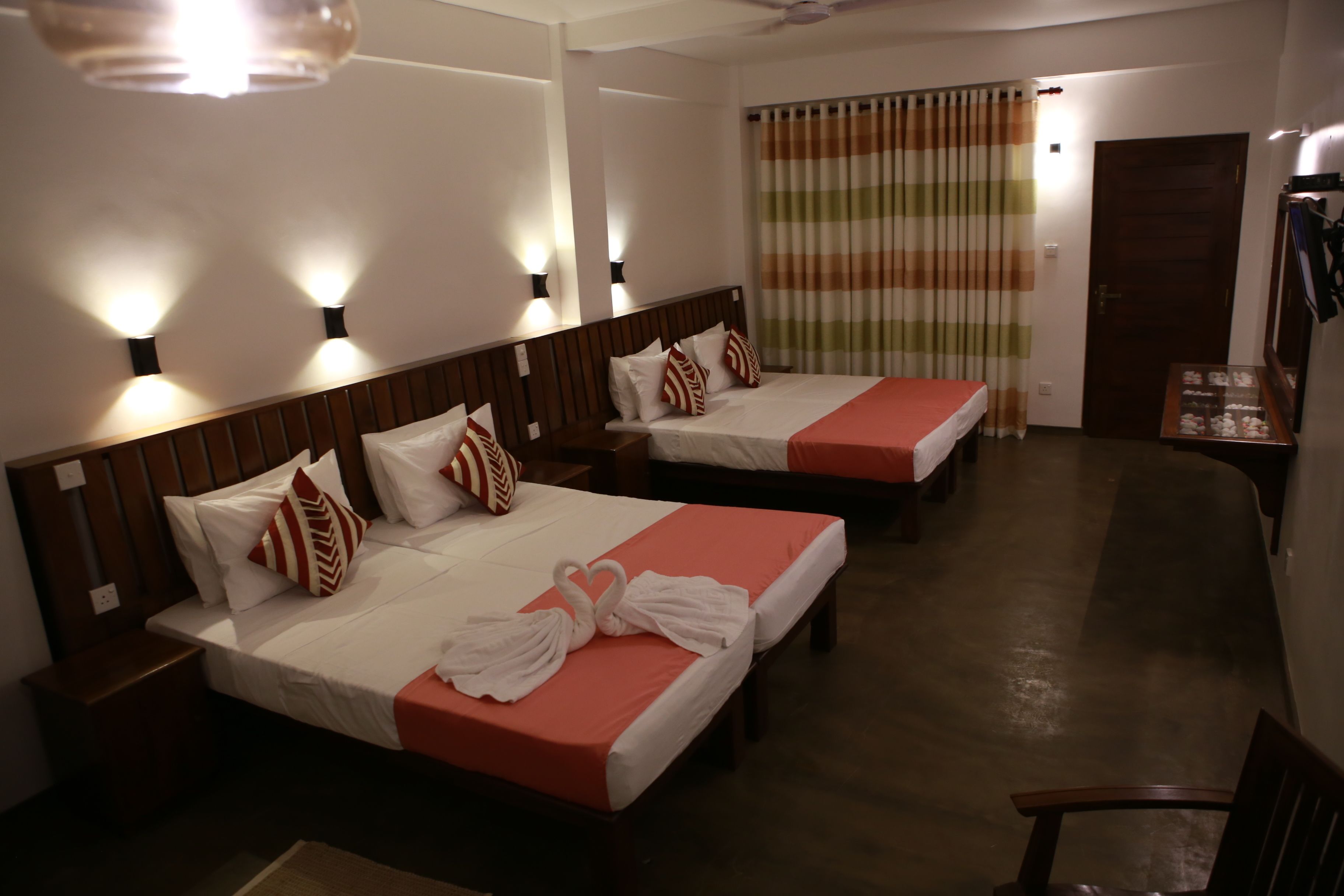 A hotel room with two beds, each with a red bedspread and white pillows, and a wooden headboard. There are wall-mounted lights above each bed and a ceiling fan.