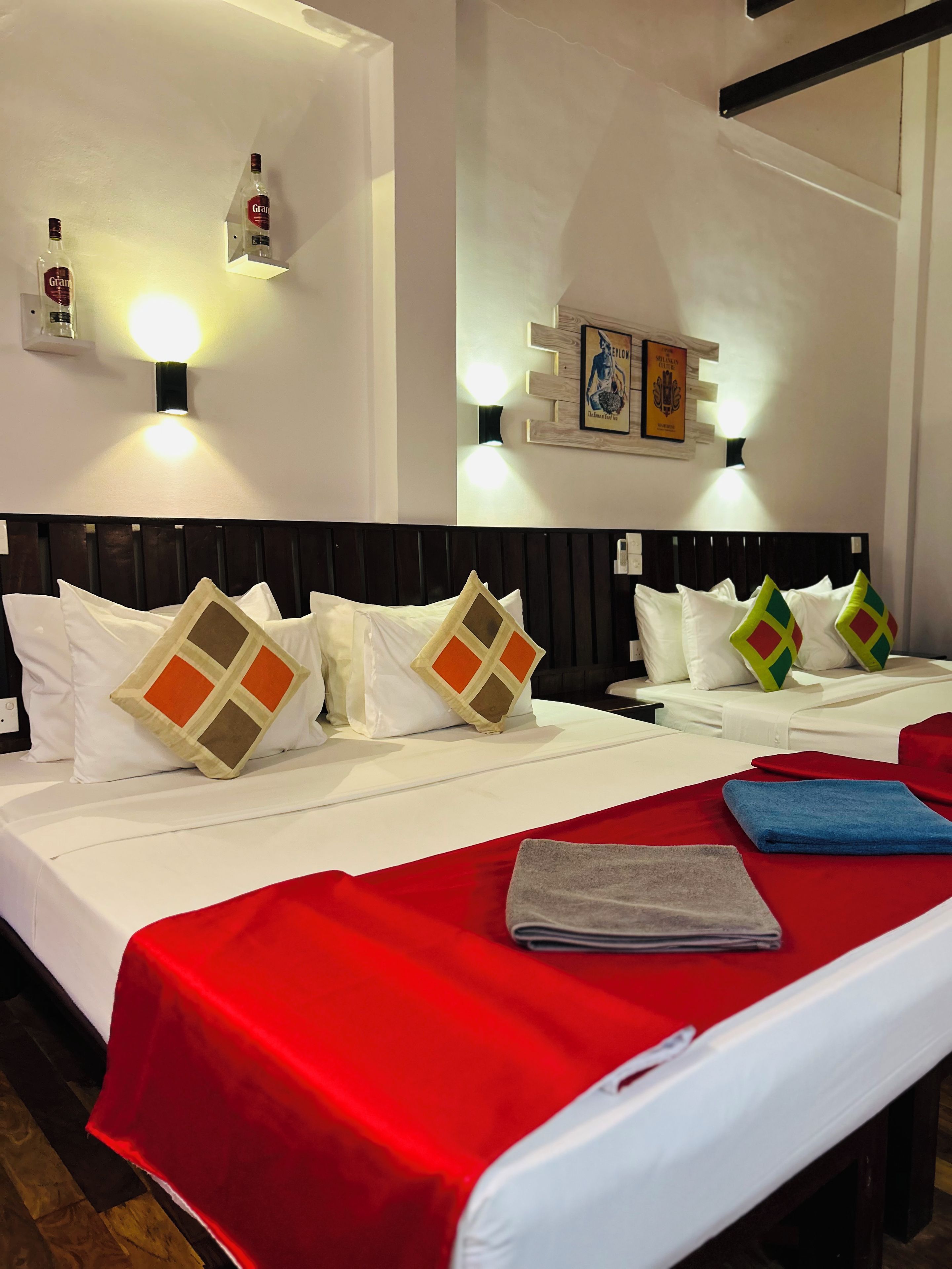 A hotel room with two beds, each with a headboard, white bedding, and decorative pillows. There are wall-mounted light fixtures and a framed picture on the wall.