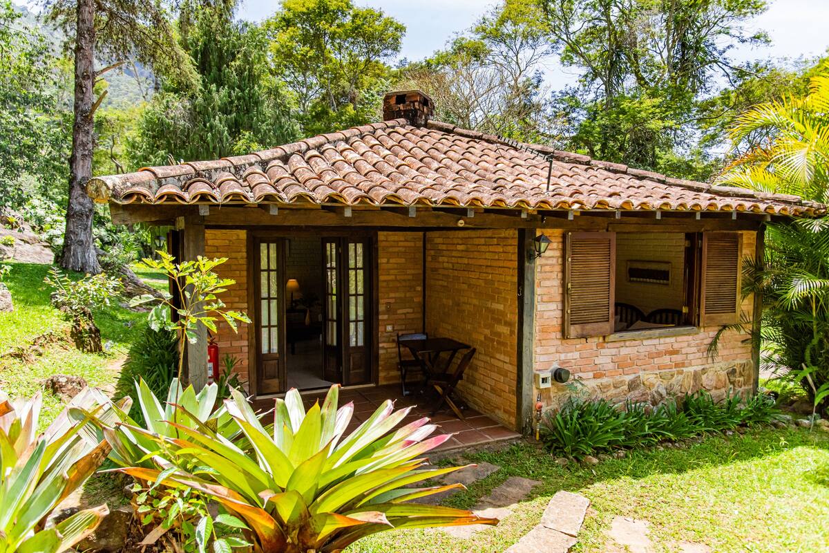 Una pequeña casa de ladrillo con tejado de baldosas, un patio con mesa y sillas, y abundante vegetación a su alrededor.