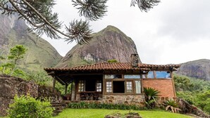 A rustic cabin with a tiled roof, stone walls, and a wooden porch. It is surrounded by lush greenery and a prominent mountain in the background. - Pousada das Araras (Petrópolis)